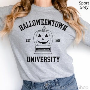 Puede incluir: Una sudadera gris con el texto "HALLOWEENTOWN UNIVERSITY" en negro. El texto est&aacute; en una fuente de estilo vintage. Hay una ilustraci&oacute;n en blanco y negro de una calabaza con una sonrisa de dientes y el texto "EST. 1998" debajo de la calabaza.
