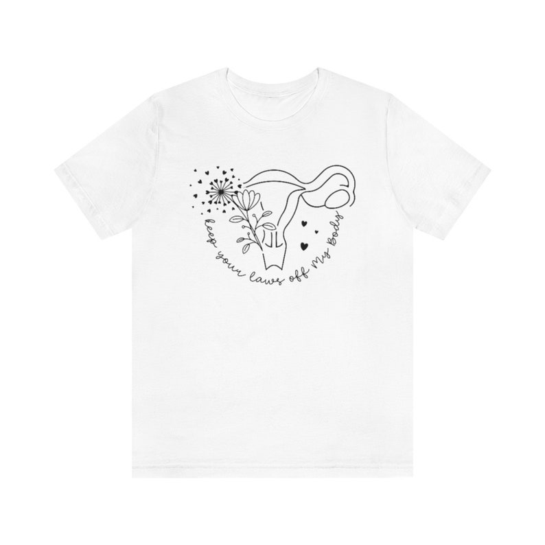Peut inclure: T-shirt blanc avec un dessin au trait noir d'un ut&eacute;rus avec une fleur et des feuilles &agrave; l'int&eacute;rieur. Le texte "keep your laws off my body" est &eacute;crit en cercle autour de l'ut&eacute;rus.