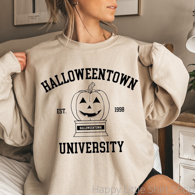 Puede incluir: Una sudadera beige con un estampado gr&aacute;fico negro de una calabaza con una cara sonriente. El texto "HALLOWEENTOWN UNIVERSITY EST. 1998" est&aacute; impreso en la sudadera.