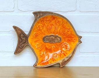 May include: A ceramic fish-shaped dish with a vibrant orange and yellow interior. The dish has a brown border and tail fins, with the word "HAWAII" in the center. The design includes floral and seahorse details, perfect for serving snacks or as a decorative piece.