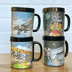 May include: Four tall, cylindrical mugs with black handles and rims. The mugs feature colorful illustrations of vintage airplanes, a lunar module, and a space shuttle. The mugs are stacked on a wooden surface.