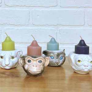 May include: Four ceramic candle holders shaped like animal faces: a lion, monkey, cat, and dog. Each holder has a different colored candle: green, brown, blue, and purple. The holders are white, with painted details.