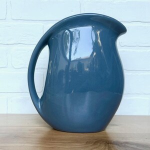 May include: A blue ceramic pitcher with a smooth, glossy finish. The pitcher has a wide, rounded base and a narrow spout.