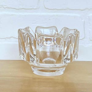 May include: A clear, faceted glass bowl with a unique, scalloped rim. The bowl has a square base and a central well. The glass is transparent, allowing light to pass through. The bowl is likely used for decorative purposes or as a small dish.