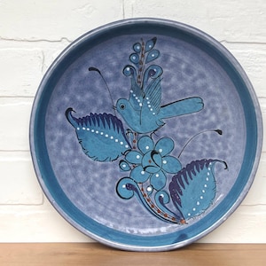 May include: A blue ceramic plate with a hand-painted design of a bird and flowers. The plate has a light blue background with dark blue accents.