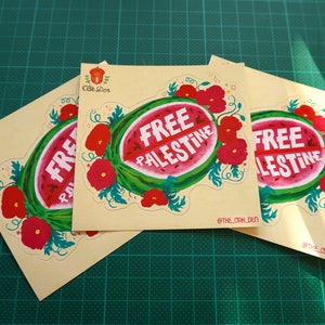 Peut inclure: Trois autocollants carr&eacute;s avec un motif de past&egrave;que et l'inscription blanche "FREE PALESTINE". La past&egrave;que est entour&eacute;e de fleurs rouges et de feuilles vertes. Les autocollants sont sur un tapis de coupe vert.