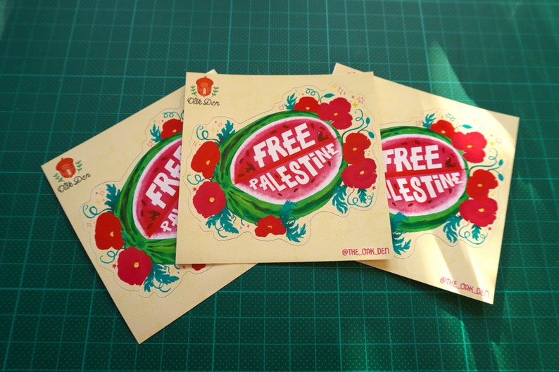 Peut inclure: Trois autocollants carr&eacute;s avec un motif de past&egrave;que et l'inscription blanche "FREE PALESTINE". La past&egrave;que est entour&eacute;e de fleurs rouges et de feuilles vertes. Les autocollants sont sur un tapis de coupe vert.
