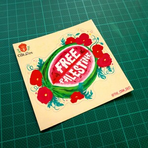 Peut inclure: Un autocollant carr&eacute; avec un motif de past&egrave;que et les mots "FREE PALESTINE" en blanc. La past&egrave;que est entour&eacute;e de fleurs rouges et de feuilles vertes. L'autocollant est sur un tapis de coupe vert.