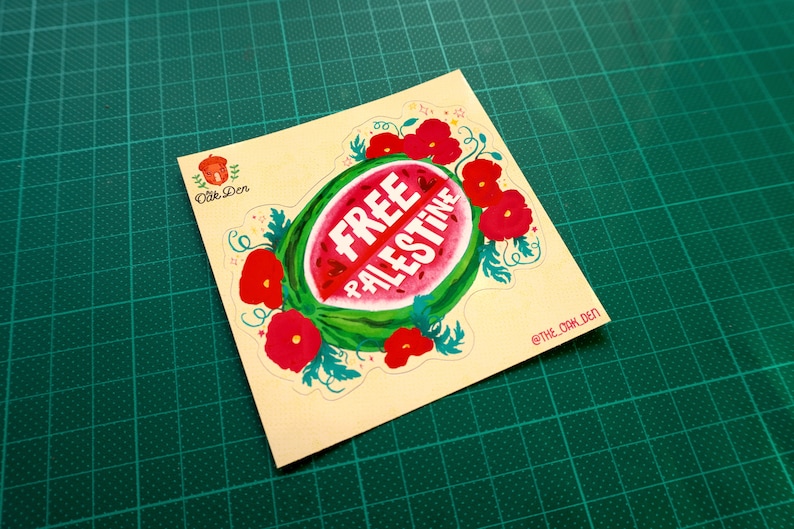 Peut inclure: Un autocollant carr&eacute; avec un motif de past&egrave;que et les mots "FREE PALESTINE" en blanc. La past&egrave;que est entour&eacute;e de fleurs rouges et de feuilles vertes. L'autocollant est sur un tapis de coupe vert.