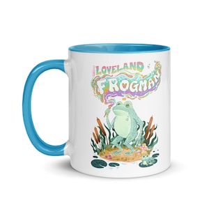 Puede incluir: Taza de cerámica blanca con interior y asa azules. La taza presenta una ilustración caprichosa de una rana con corona, sosteniendo una varita mágica, y el texto "The Loveland Frogman" en letras coloridas.