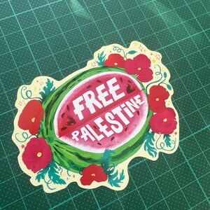 Peut inclure: Un autocollant d&eacute;coup&eacute; repr&eacute;sentant une tranche de past&egrave;que avec l'inscription "FREE PALESTINE" en blanc. La past&egrave;que est entour&eacute;e de coquelicots rouges et de feuilles vertes, sur un fond jaune clair. L'autocollant est pos&eacute; sur un tapis de d&eacute;coupe vert.