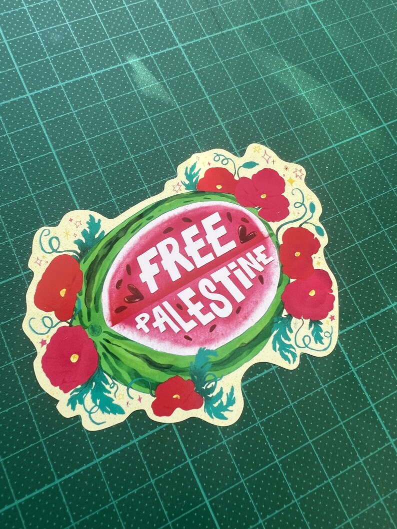 Peut inclure: Un autocollant d&eacute;coup&eacute; repr&eacute;sentant une tranche de past&egrave;que avec l'inscription "FREE PALESTINE" en blanc. La past&egrave;que est entour&eacute;e de coquelicots rouges et de feuilles vertes, sur un fond jaune clair. L'autocollant est pos&eacute; sur un tapis de d&eacute;coupe vert.