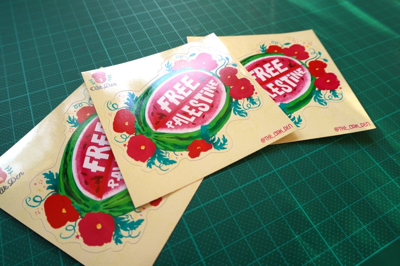 Peut inclure: Trois autocollants avec un motif de past&egrave;que et les mots "FREE PALESTINE" en blanc. La past&egrave;que est entour&eacute;e de fleurs rouges et de feuilles vertes. Les autocollants sont sur un fond beige clair.