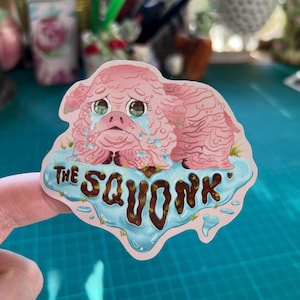 Könnte beinhalten: Ein rosa Aufkleber mit einer traurigen, weinenden Kreatur mit dem Text "THE SAVONK" in braunen Buchstaben. Die Kreatur ist in einer blauen Wasserpfütze dargestellt.