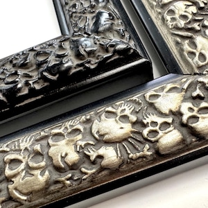May include: Two black picture frames with ornate silver skull designs. The frames are made of wood and have a black finish. The skull designs are raised and detailed.
