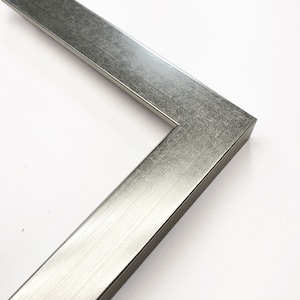 Op de afbeelding: Close-up van een hoek van een zilveren metalen fotolijst. De lijst heeft een geborstelde metalen afwerking en een rechthoekig profiel. De hoek is in een hoek van 90 graden verbonden, wat de constructie van de lijst laat zien. Geschikt voor kunstwerken of foto's.