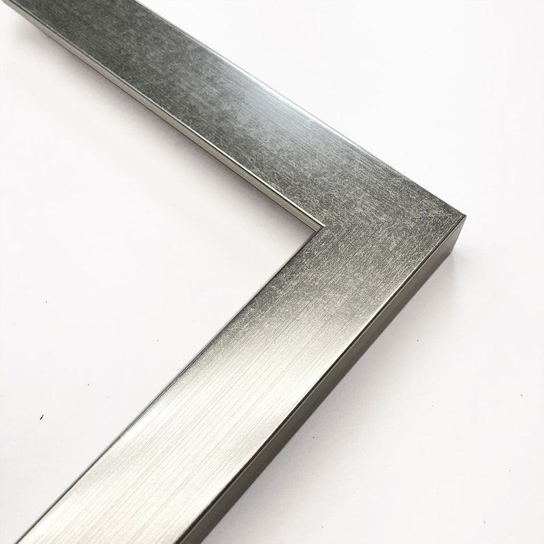 Op de afbeelding: Close-up van een hoek van een zilveren metalen fotolijst. De lijst heeft een geborstelde metalen afwerking en een rechthoekig profiel. De hoek is in een hoek van 90 graden verbonden, wat de constructie van de lijst laat zien. Geschikt voor kunstwerken of foto's.