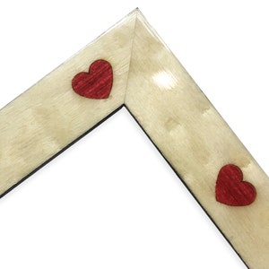 May include: A corner of a wooden picture frame with a light beige finish. Two red heart-shaped accents are placed on the frame. The frame has a black interior edge and is set against a white background.