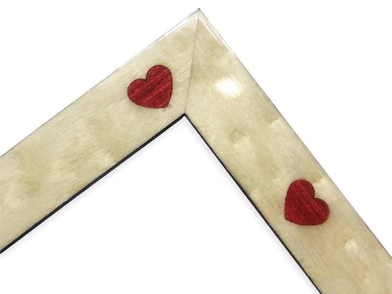 May include: A corner of a wooden picture frame with a light beige finish. Two red heart-shaped accents are placed on the frame. The frame has a black interior edge and is set against a white background.