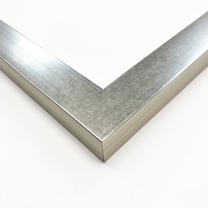 Op de afbeelding: Close-up van een zilveren hoek van een fotolijst. Het frame heeft een geborstelde metalen textuur en een strak, modern design. De hoek is in een hoek van 45 graden verbonden, wat de constructie van het frame laat zien. De achtergrond is wit.