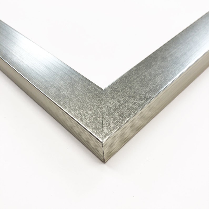 Op de afbeelding: Close-up van een zilveren hoek van een fotolijst. Het frame heeft een geborstelde metalen textuur en een strak, modern design. De hoek is in een hoek van 45 graden verbonden, wat de constructie van het frame laat zien. De achtergrond is wit.