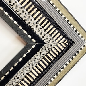 May include: Close-up of a corner of a decorative picture frame with a multi-layered design. The frame features a black, white, and gray geometric pattern.