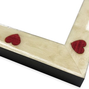 May include: Close-up of a picture frame corner. The frame has a light wood grain finish with small, red heart-shaped accents. The frame's edge is black, creating a contrast with the lighter wood and red hearts. The frame is designed for displaying photos.