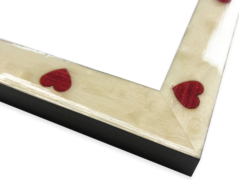 May include: Close-up of a picture frame corner. The frame has a light wood grain finish with small, red heart-shaped accents. The frame's edge is black, creating a contrast with the lighter wood and red hearts. The frame is designed for displaying photos.