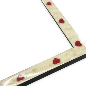 May include: A corner of a picture frame with a cream-colored surface and red heart-shaped accents. The frame has a black edge and a light-colored, textured surface. The hearts are evenly spaced.