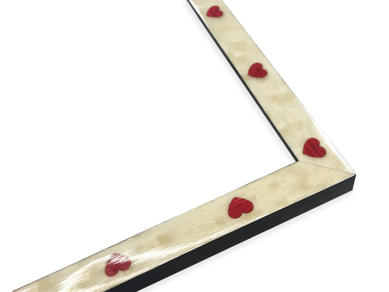 May include: A corner of a picture frame with a cream-colored surface and red heart-shaped accents. The frame has a black edge and a light-colored, textured surface. The hearts are evenly spaced.