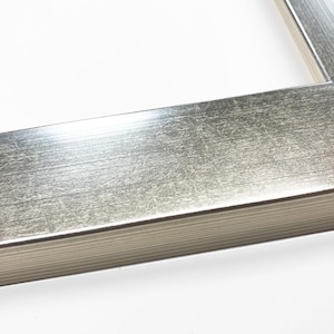 Op de afbeelding: Close-up van een zilverkleurige fotolijst met een getextureerd oppervlak. De lijst heeft een rechthoekige vorm en een metallic afwerking. De lijst staat tegen een witte achtergrond.