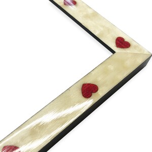 May include: Angled view of a picture frame corner. The frame has a light beige, textured surface with small, red heart-shaped embellishments. The frame's edges are black, and the background is white.