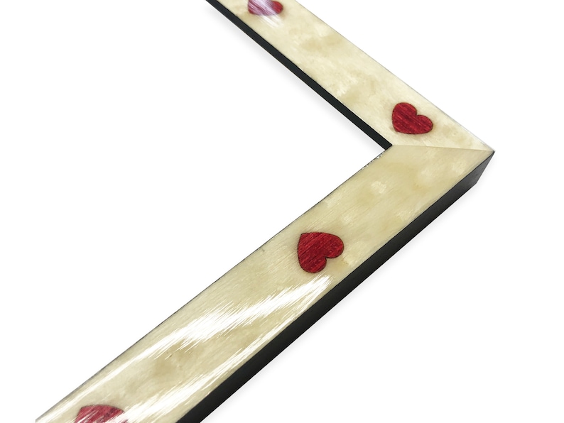 May include: Angled view of a picture frame corner. The frame has a light beige, textured surface with small, red heart-shaped embellishments. The frame's edges are black, and the background is white.
