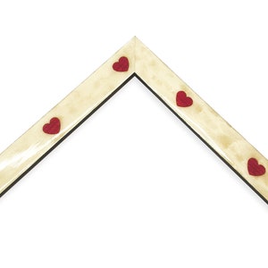 May include: A corner of a picture frame with a light beige finish and a black interior edge. Small red heart-shaped embellishments are evenly spaced along the frame's edge. The frame is set against a white background.