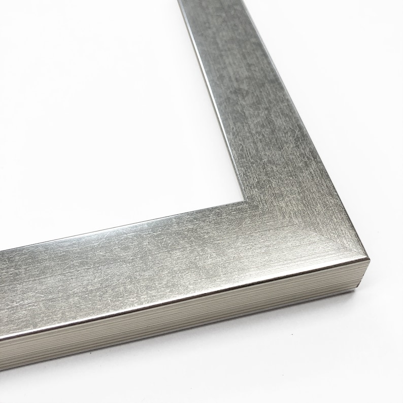 Op de afbeelding: Een zilverkleurige fotolijst met een getextureerde, geborstelde metalen afwerking. De lijst heeft een strak, modern ontwerp met een afgeschuinde rand en een wit interieur. De hoek van de lijst is zichtbaar en toont het precieze vakmanschap.