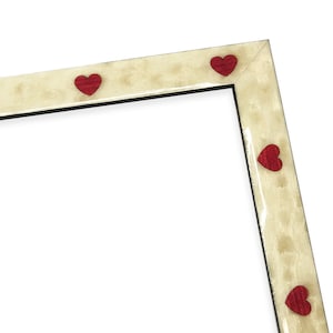 May include: A cream-colored picture frame with red heart-shaped accents. The frame has a textured finish and a black interior border. The hearts are evenly spaced along the frame's edges, creating a decorative border.