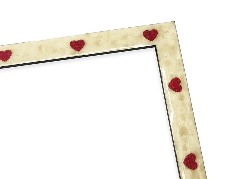 May include: A cream-colored picture frame with red heart-shaped accents. The frame has a textured finish and a black interior border. The hearts are evenly spaced along the frame's edges, creating a decorative border.