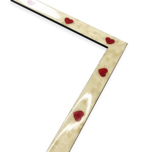 May include: Angled wooden picture frame with a light beige finish and black edges. The frame is adorned with small, red heart-shaped embellishments. The design is simple and elegant, suitable for displaying photographs or artwork.
