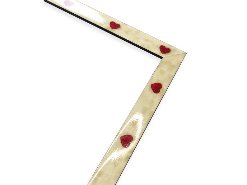 May include: Angled wooden picture frame with a light beige finish and black edges. The frame is adorned with small, red heart-shaped embellishments. The design is simple and elegant, suitable for displaying photographs or artwork.