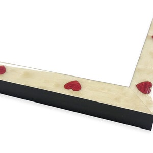 May include: A black picture frame with a cream-colored border. The border is decorated with evenly spaced red heart-shaped accents. The frame is angled, showing the corner and a portion of the interior.