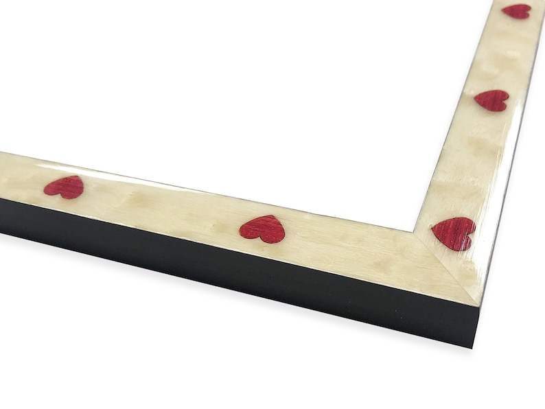 May include: A black picture frame with a cream-colored border. The border is decorated with evenly spaced red heart-shaped accents. The frame is angled, showing the corner and a portion of the interior.
