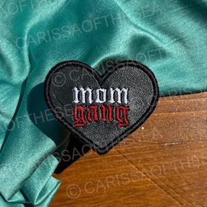 May include: A black heart-shaped patch with the words "MOM GANG" embroidered in white and red. The patch is on a wooden surface, with a teal satin fabric in the background.
