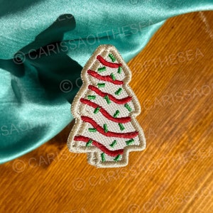 May include: An embroidered Christmas tree patch with a cream-colored base, red ribbon-like stripes, and green accents. The patch is on a wooden surface with a teal fabric in the background.