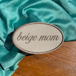 May include: An oval-shaped patch with the words "beige mom" embroidered in a cursive font. The patch has a beige background and a brown border. The patch is set against a teal satin fabric.