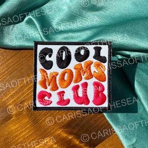 May include: A square patch with the words "COOL MOMS CLUB" in retro-style lettering. The text is in black, orange, and red, with a white background and a black border. The patch is on a wooden surface with a teal fabric.