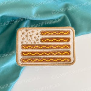 May include: A rectangular embroidered patch featuring a stylized American flag design where the stripes are replaced by brown hot dogs topped with yellow mustard lines and the blue canton is replaced by a field of small embroidered stars.