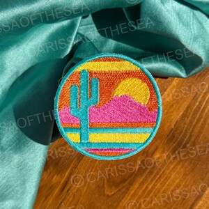 May include: A round embroidered patch with a desert sunset scene. The patch features a cactus, a pink mountain, a yellow sun, and a blue sky with orange and yellow stripes.