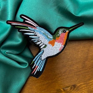 May include: A colorful embroidered hummingbird patch with a teal blue, pink, orange, and green design. The patch is on a teal blue fabric background.