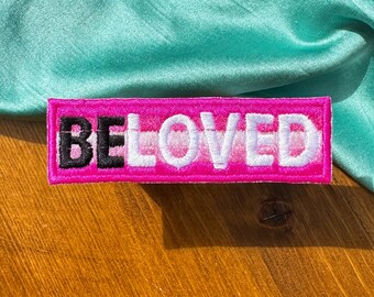 May include: A pink and white embroidered patch with the word "BELOVED" in white letters on a pink background. The patch has a black border.