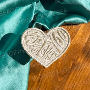May include: White heart-shaped patch with the words "Just Love" embroidered in a cursive font. The patch is on a wooden surface.